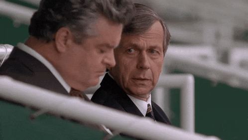 a man in a suit and tie sits next to another man in a stadium