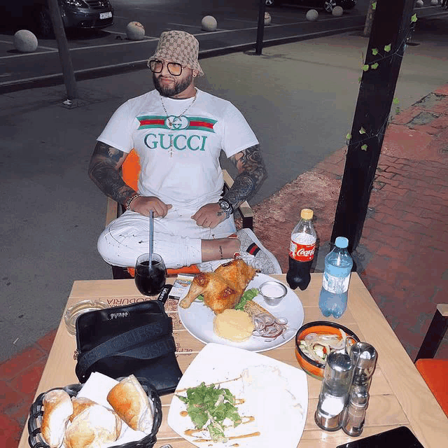 a man wearing a white gucci shirt sits at a table with food