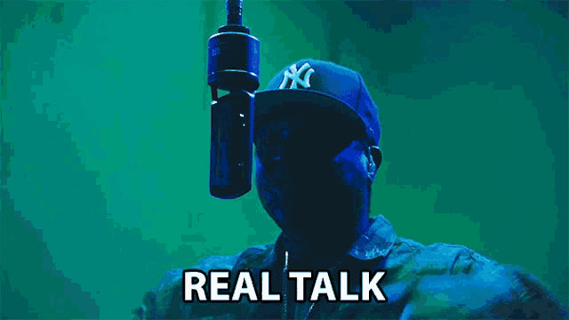 a man wearing a ny hat stands in front of a microphone with the words real talk written below him