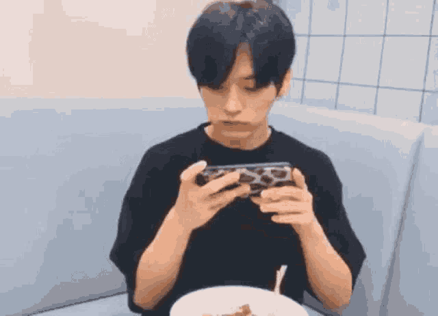 a young man is sitting on a couch eating noodles and playing a game on his phone .