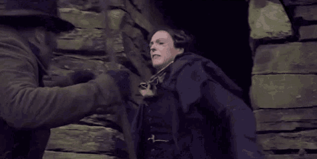 a man in a cape is standing next to a brick wall and fighting another man .