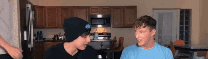 two young men are sitting in a kitchen and talking to each other .