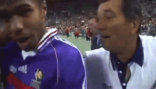 two men are standing next to each other on a soccer field . one of the men is wearing a blue jersey .