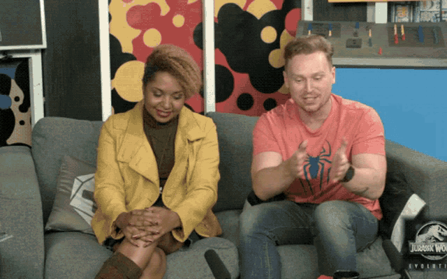a man and a woman sit on a couch with a jurassic world evolution blanket