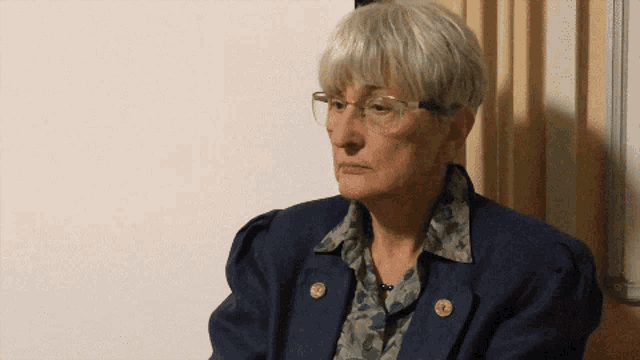 a woman wearing glasses and a blue jacket is sitting in front of a wall