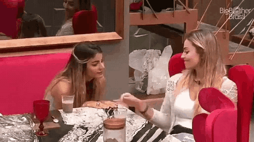two women are sitting at a table talking to each other while a mirror is behind them .