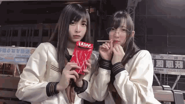 two girls in school uniforms are sitting on a bench eating a snack .