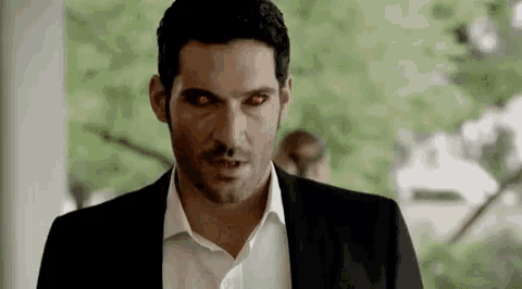 a man in a suit and white shirt with red eyes is standing in front of a tree .