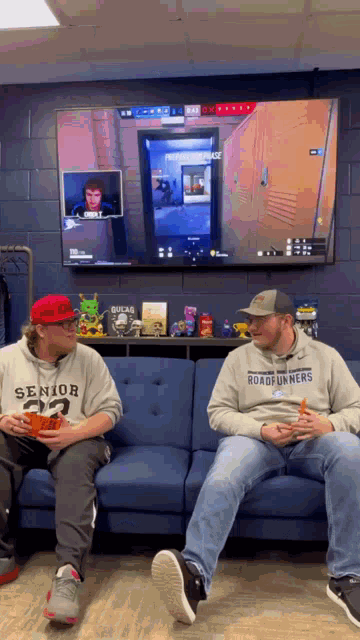 two men sitting on a couch playing a video game with one wearing a sweatshirt that says senior 23