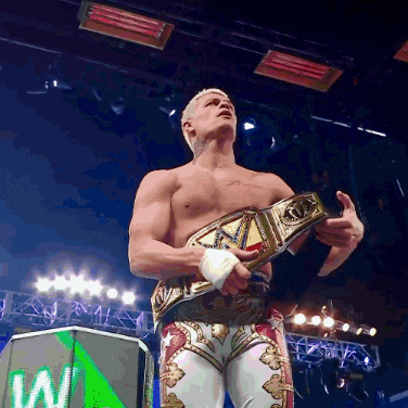 a shirtless wrestler holds a wrestling belt in front of a sign that says w