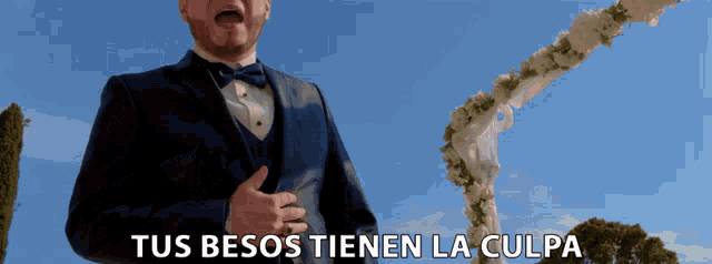 a man in a tuxedo is standing in front of a wedding arch and says tus besos tienen la culpa