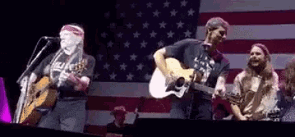 a group of men are playing guitars and singing on stage in front of an american flag .