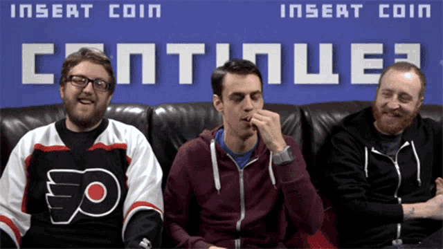 three men sit on a couch in front of a sign that says insert coin continue