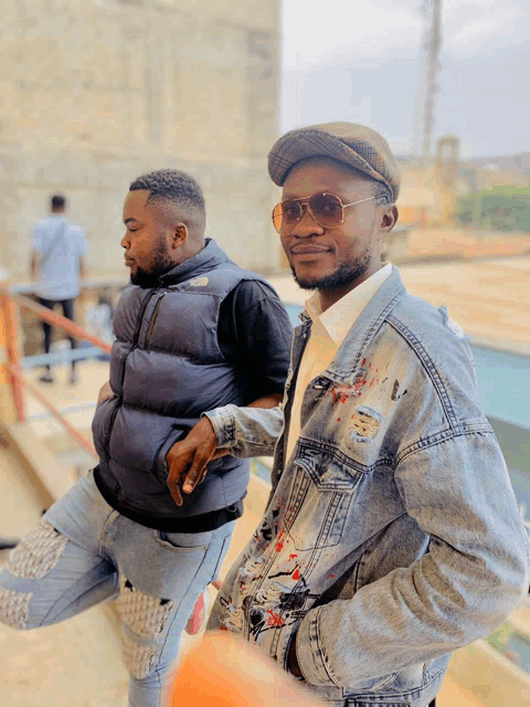 a man wearing a hat and sunglasses stands next to another man wearing a denim jacket