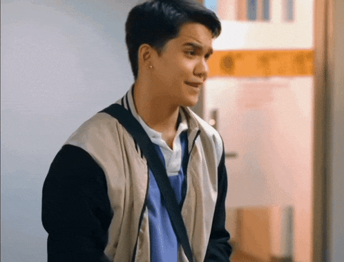 a young man standing in a hallway wearing a jacket and a blue shirt