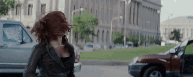 a woman with red hair is walking down a street in front of a building and a car .