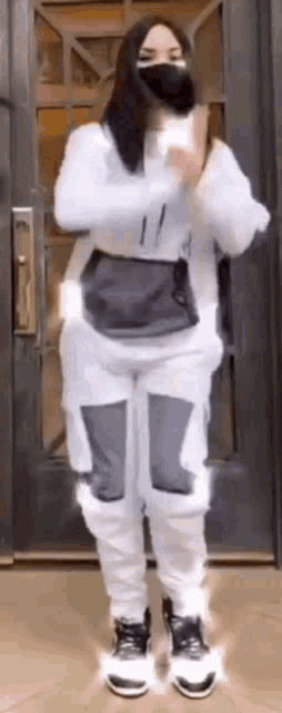 a woman wearing a mask and white sweatpants is standing in front of a door and dancing .