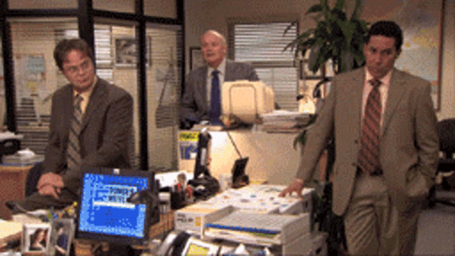 three men in suits and ties are standing around a desk with a dell computer on it