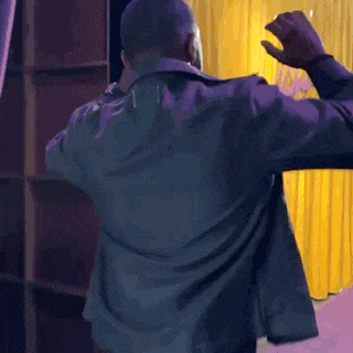 a man in a black jacket is dancing in front of a yellow curtain
