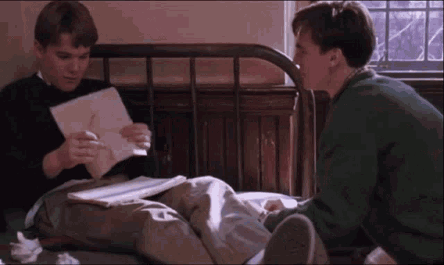 two men are sitting on a bed looking at papers