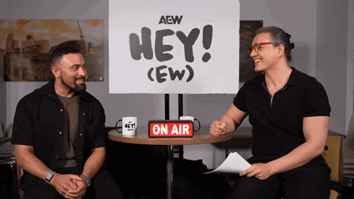 two men sit at a table in front of a sign that says hey ew