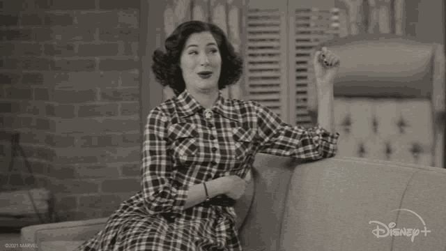 a woman in a plaid dress is sitting on a couch with a disney + logo in the corner