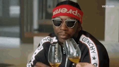 a man wearing sunglasses and a headband that says supreme is drinking from two wine glasses .