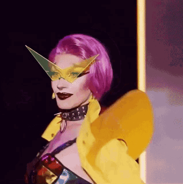 a drag queen with pink hair and a yellow jacket is wearing a mask and sunglasses .