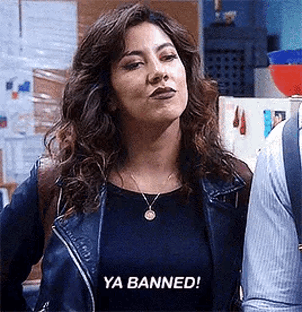 a woman wearing a leather jacket and a necklace is standing next to a man and saying `` ya banned '' .