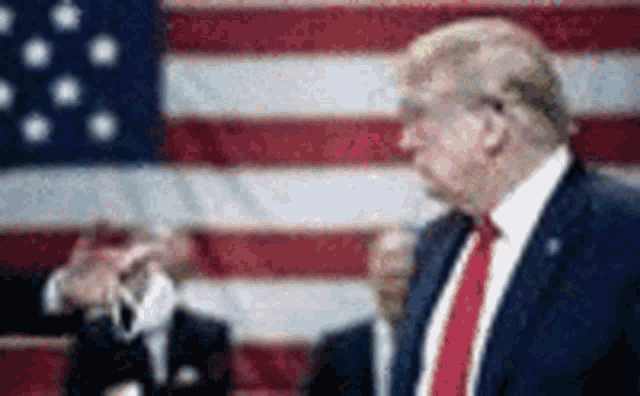 donald trump is standing in front of an american flag in a suit and tie .
