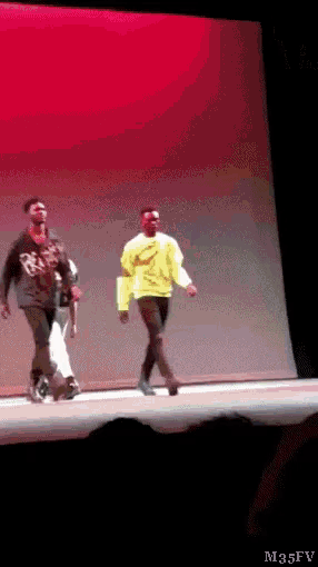 a man in a yellow sweater is walking on a stage with another man
