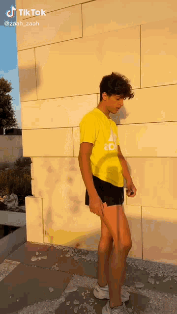a young man wearing a yellow adidas shirt and black shorts is standing in front of a wall .