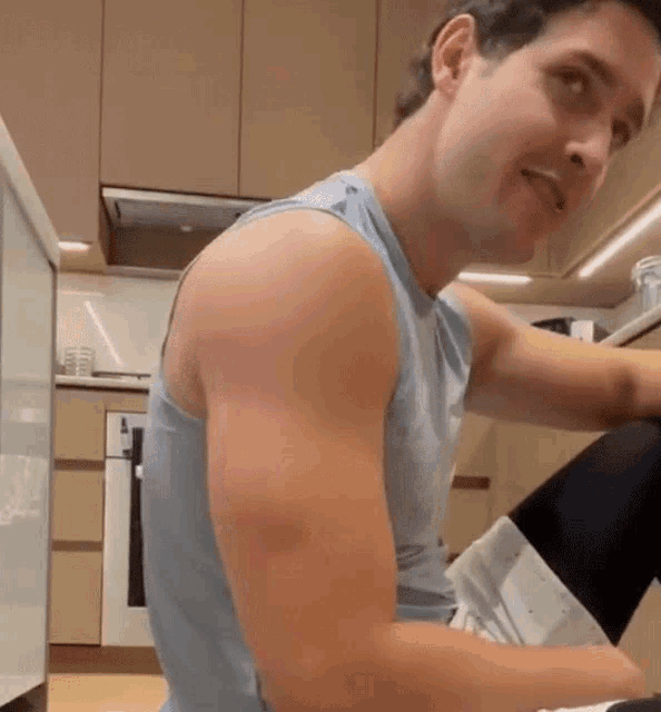 a man in a blue tank top is sitting in a kitchen looking at something .