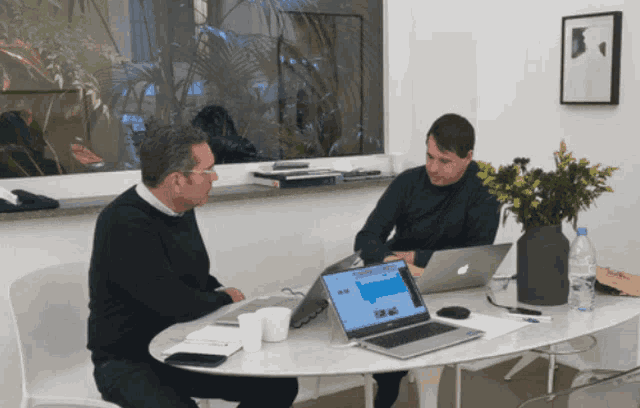 two men sit at a table with laptops and a vase of flowers on it