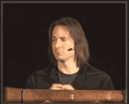 a man with long hair is sitting at a podium with a microphone
