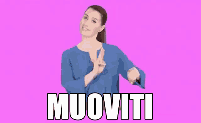 a woman in a blue shirt is standing in front of a pink background and making a gesture with her hands .