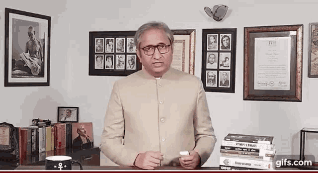 a man in a suit and glasses is standing in front of a stack of books and a framed picture of mahatma gandhi
