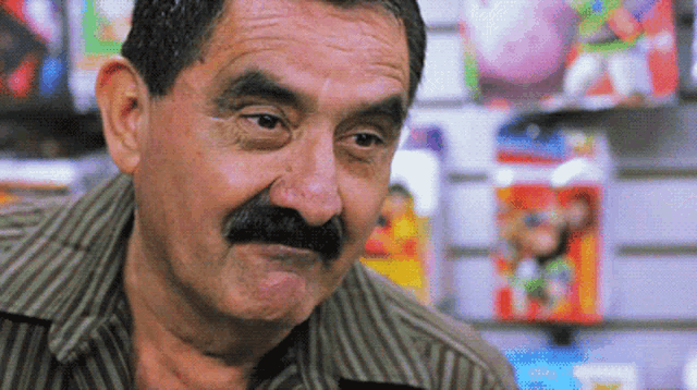 a man with a mustache looks at the camera in front of a wall of toys