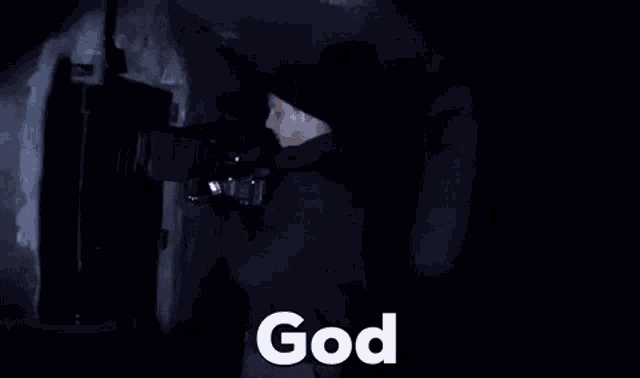 a man in a black jacket is holding a camera in a dark room and says god .