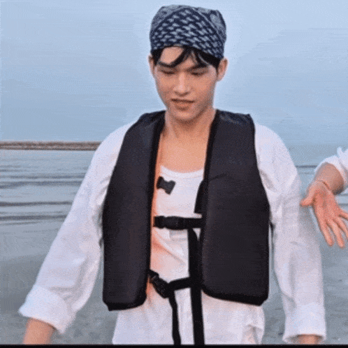 a man wearing a life jacket and a bandana stands on the beach