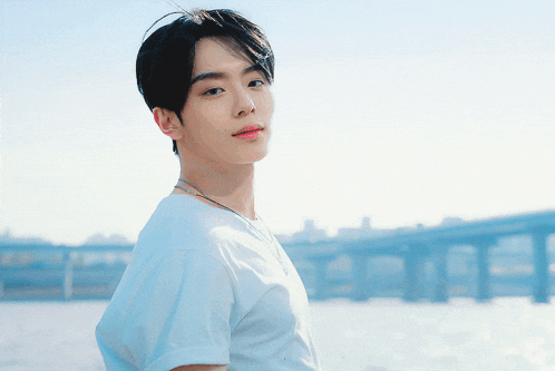 a young man in a white t-shirt stands in front of a body of water