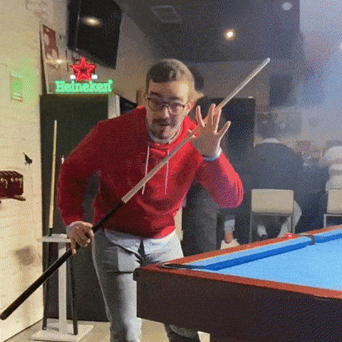 a man in a red sweatshirt is holding a pool cue in front of a heineken sign