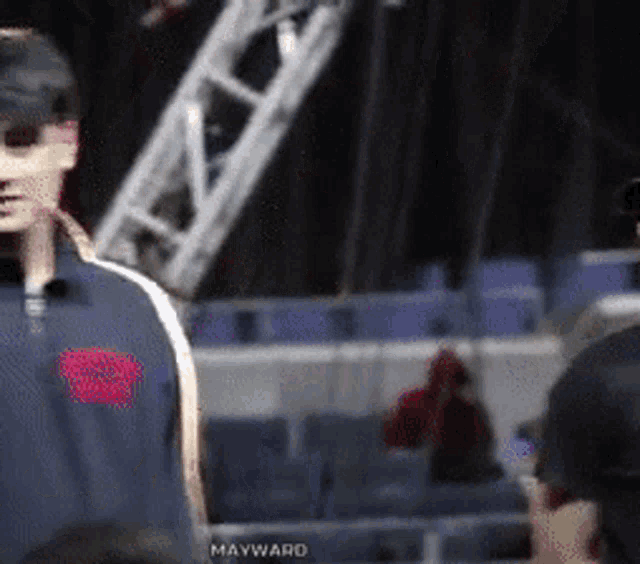 a man wearing a hat and sunglasses stands in front of a crowd with mayward written on the bottom right corner