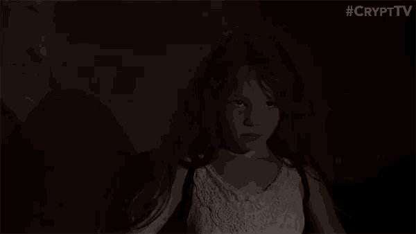 a little girl in a white dress is standing in a dark room with her arms outstretched .