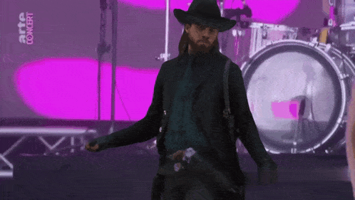 a man in a cowboy hat stands in front of a drum set with the words arte concert on the bottom right