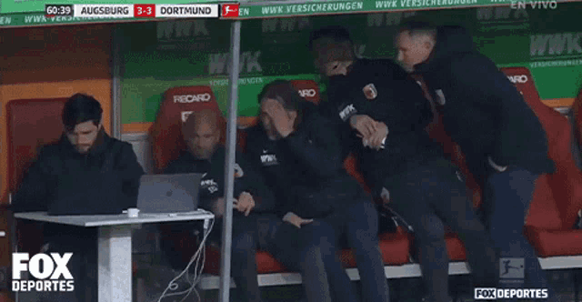 a group of men sitting in a fox deportes dugout watching a soccer game