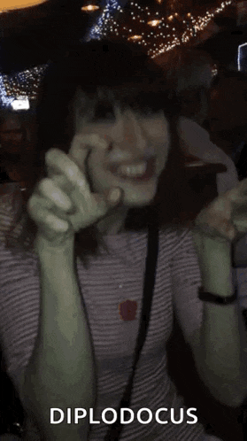 a woman in a striped shirt is smiling and dancing in a dark room with the words diplodocus written in the corner .