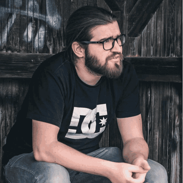 a man with glasses and a beard wears a black dc shirt
