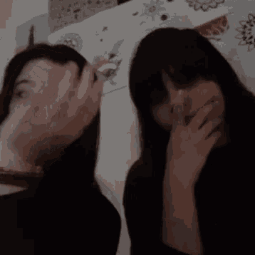 two women covering their faces with their hands in front of a wall with drawings on it