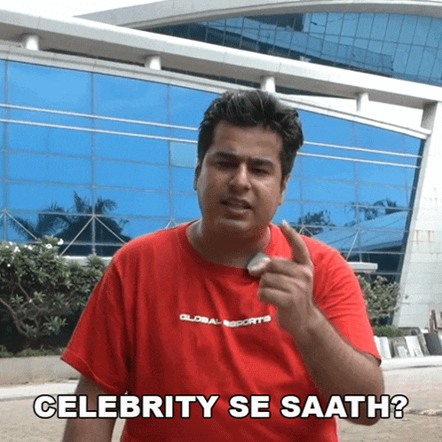 a man in a red shirt says " celebrity se saath " in front of a building